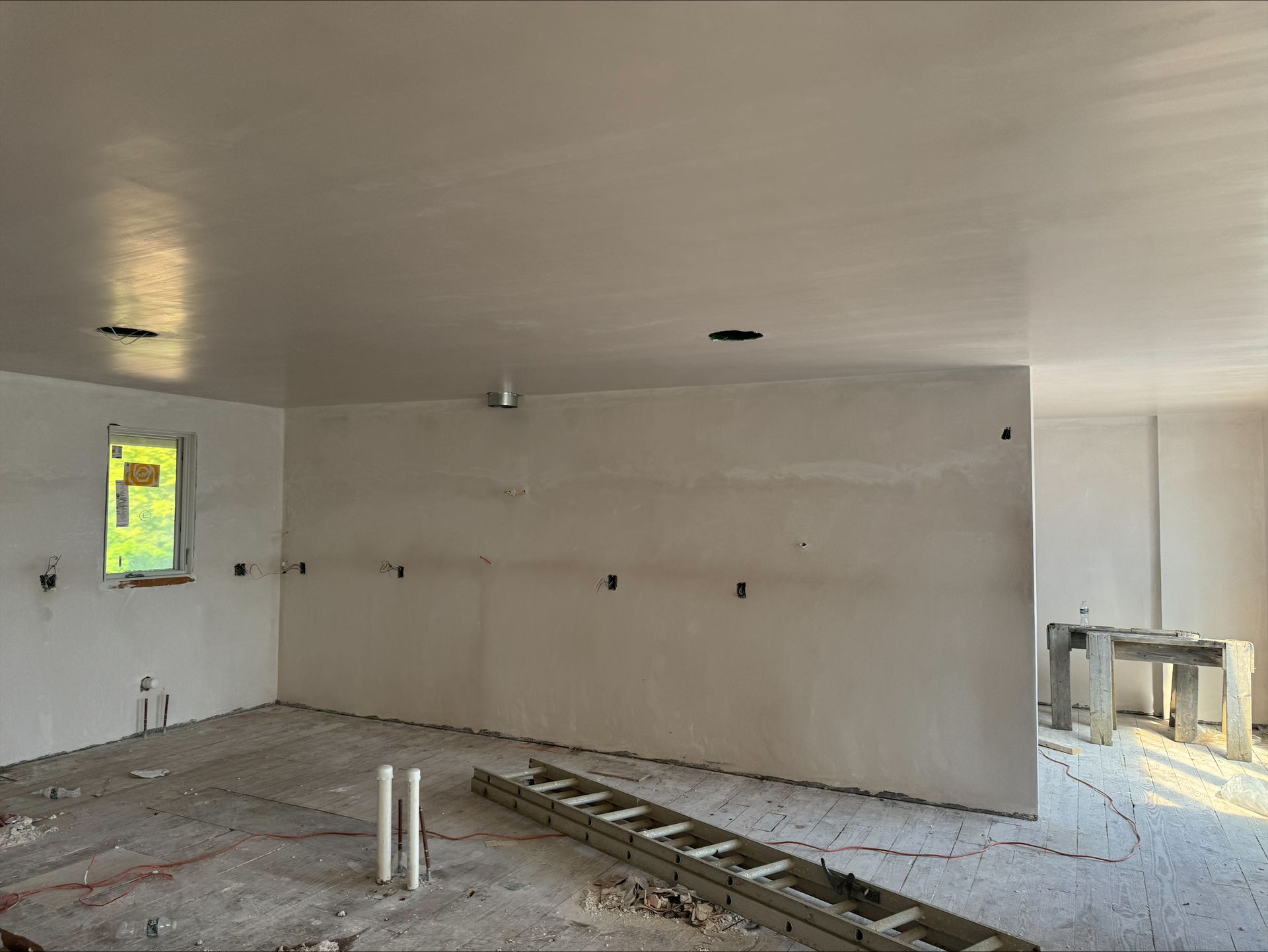 Bare interior room under construction with unfinished walls, exposed floor, and a small window on the left.