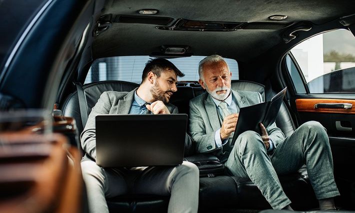 Two men are sitting in the back seat of a car looking at a laptop and a tablet.