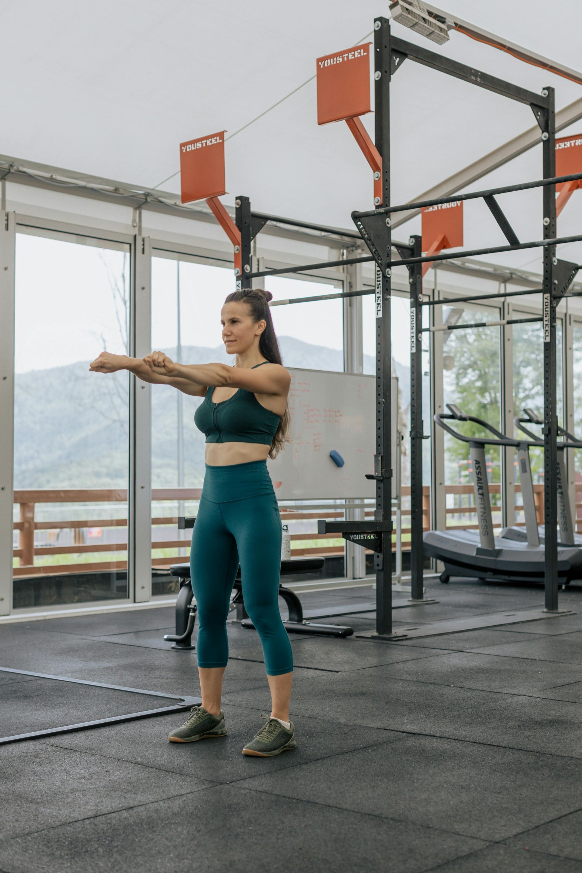 Woman in athletic wear stretches arms forward in a gym with glass walls, facing a mountain view.