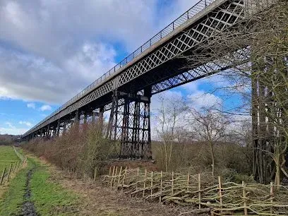 Iron railway viaduct spanning a landscape with a dirt path, green field, and bare trees under a cloudy sky.