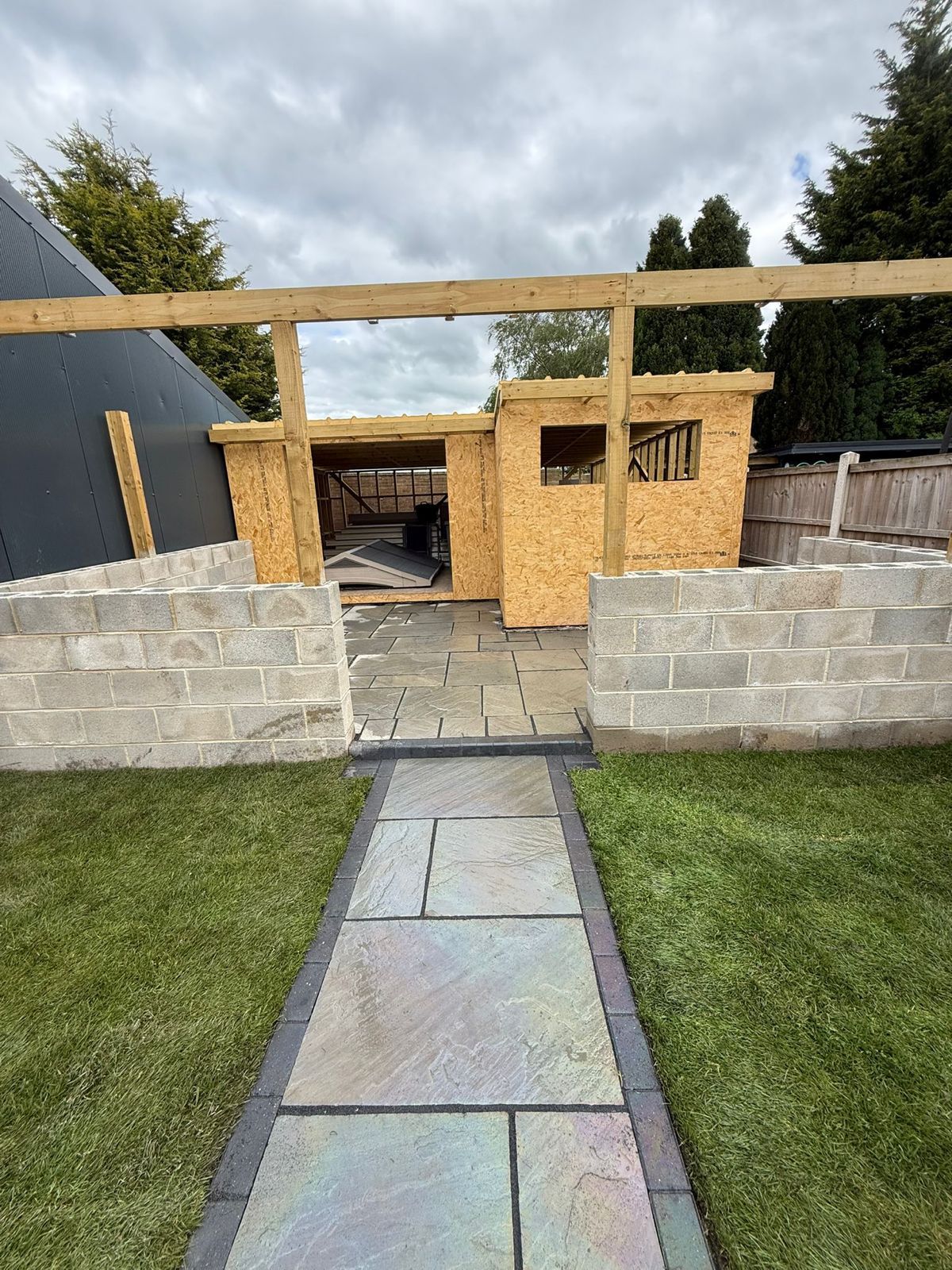 Backyard with a stone path leading to a partially built wooden shed behind a low block wall.