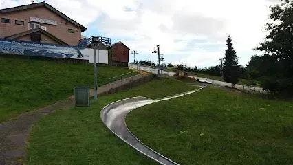 A metal toboggan run curves down a grassy hill, with a building at the top.