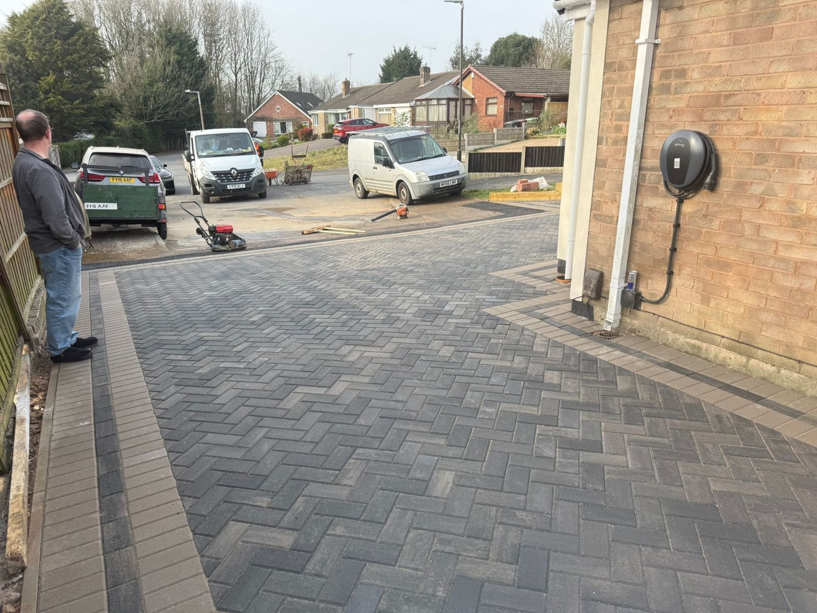 A person stands by a newly paved dark gray herringbone driveway next to a brick house with an EV charger on the wall.