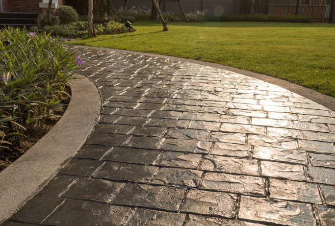 Brick pathway curves through a green lawn, edged with plants. Sunlight reflects off the pathway.