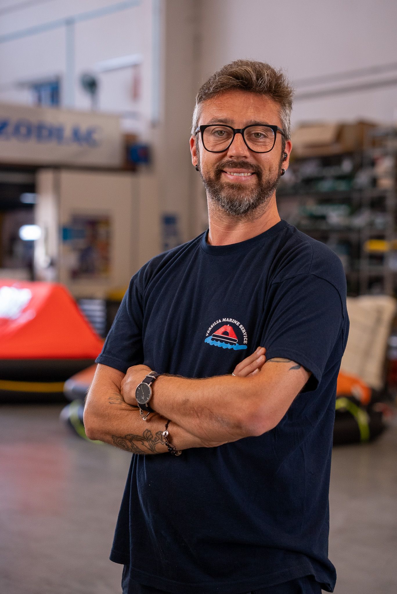 Man standing with arms crossed in a workshop, wearing a navy T-shirt and glasses, with machinery in the background