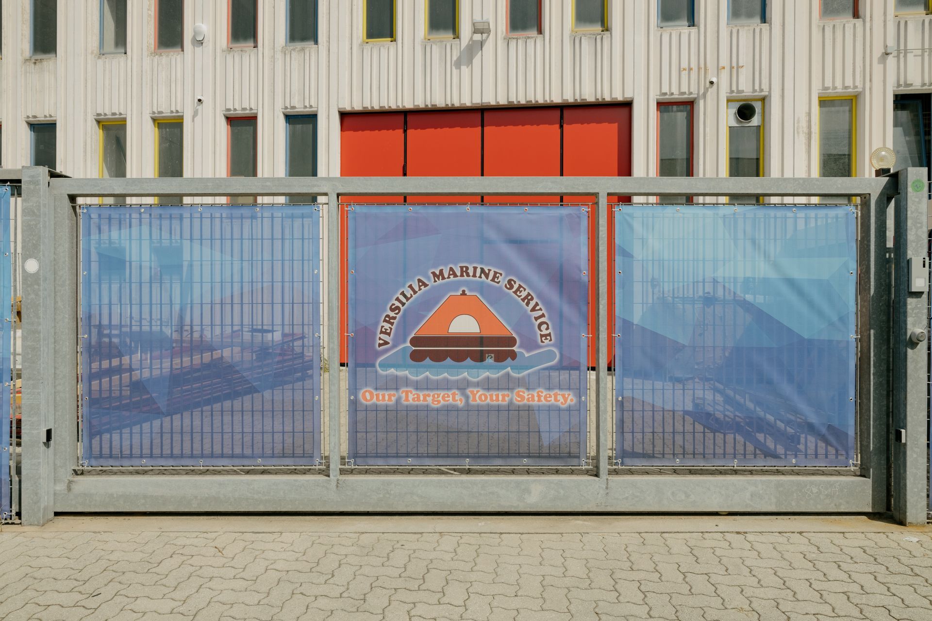 Closed metal gate with blue mesh panels and a central logo in front of an industrial building