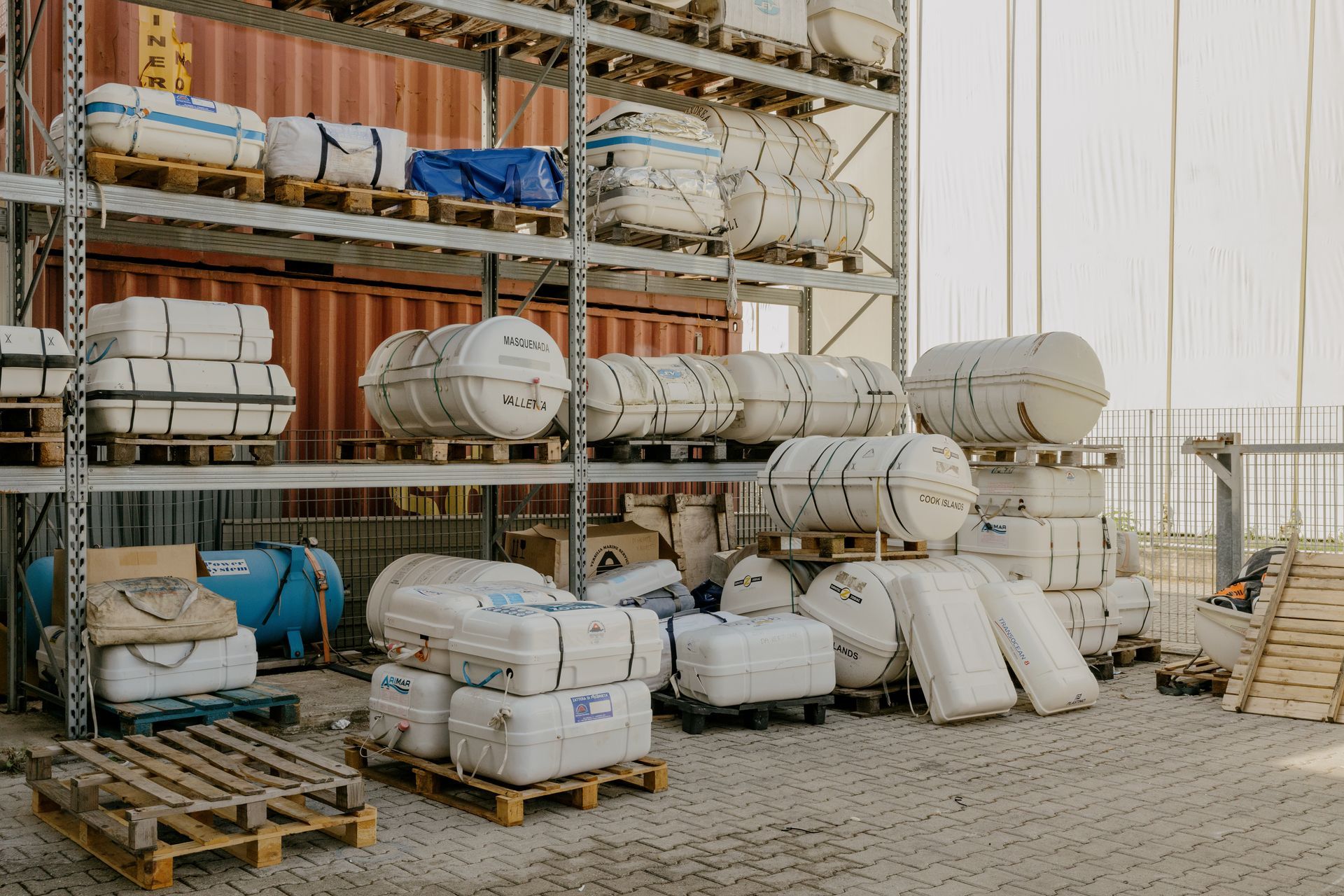 Industrial storage yard with stacked pallets of wrapped equipment and tanks beside a building