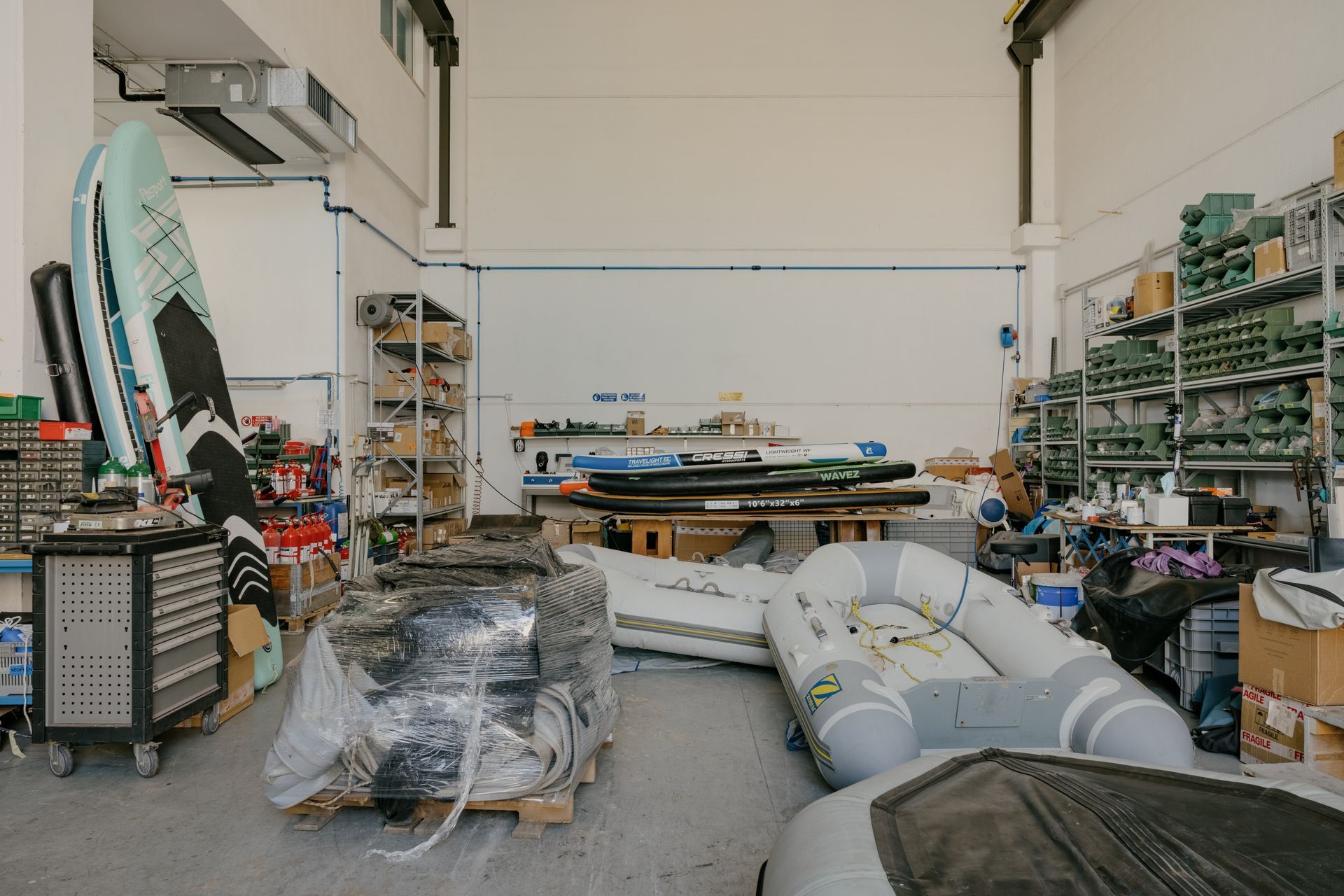 Cluttered warehouse with surfboards, stacked equipment, shelves, and large inflatable boats or kayaks.
