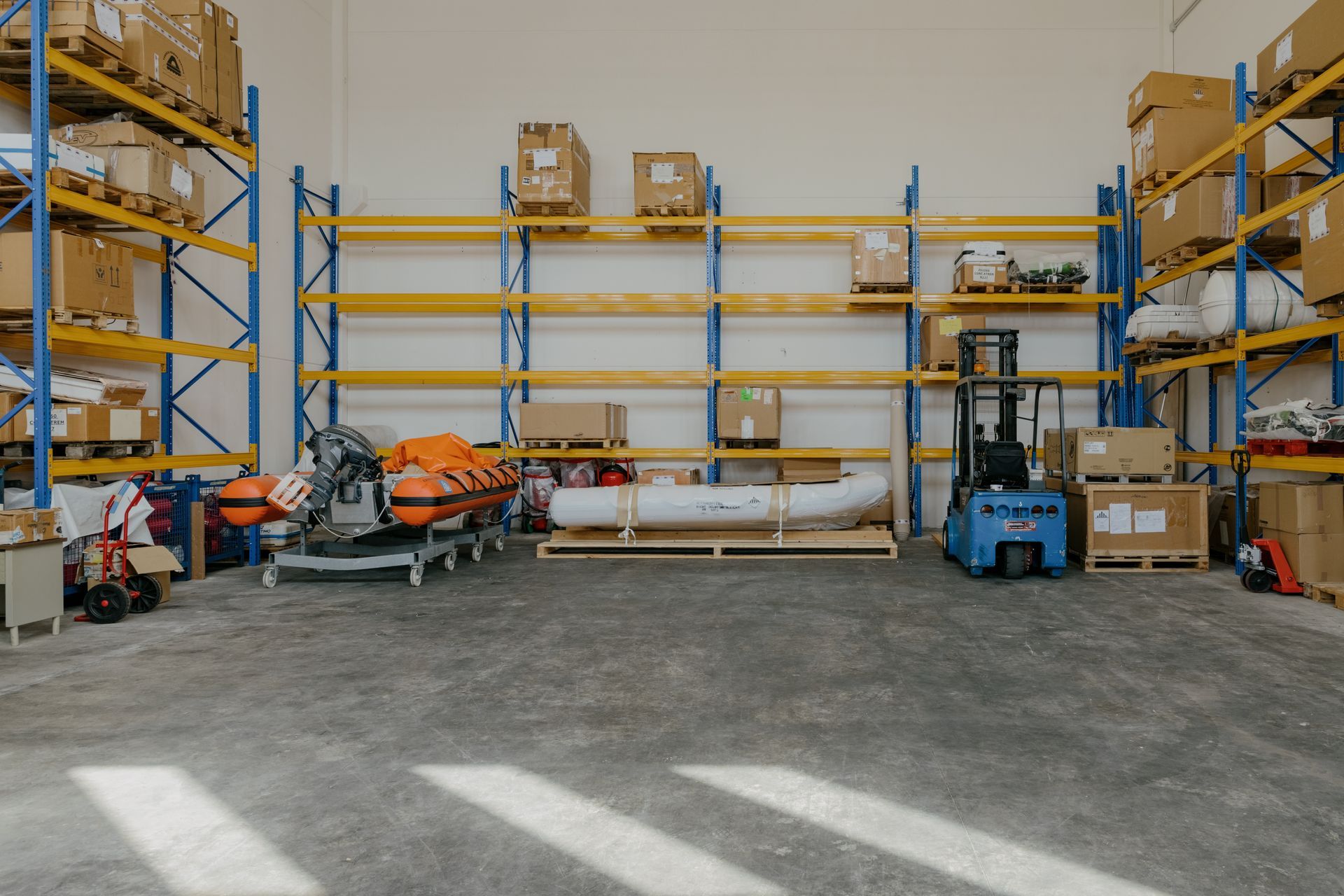 Warehouse interior with blue metal shelves, cardboard boxes, stacked orange equipment, and a forklift on a concrete floor