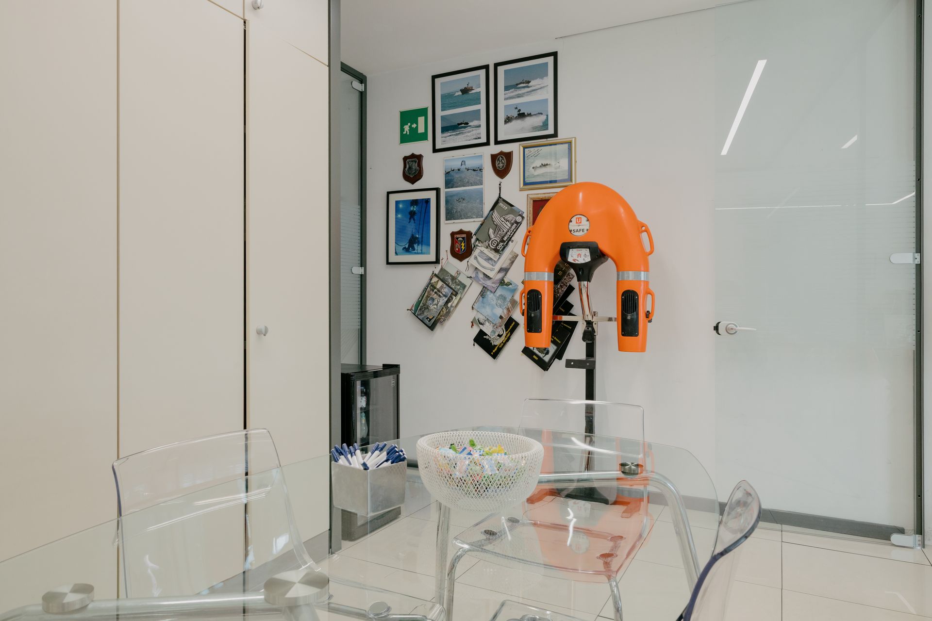 Modern office with glass conference table, orange wall-mounted device, and framed photos on the wall