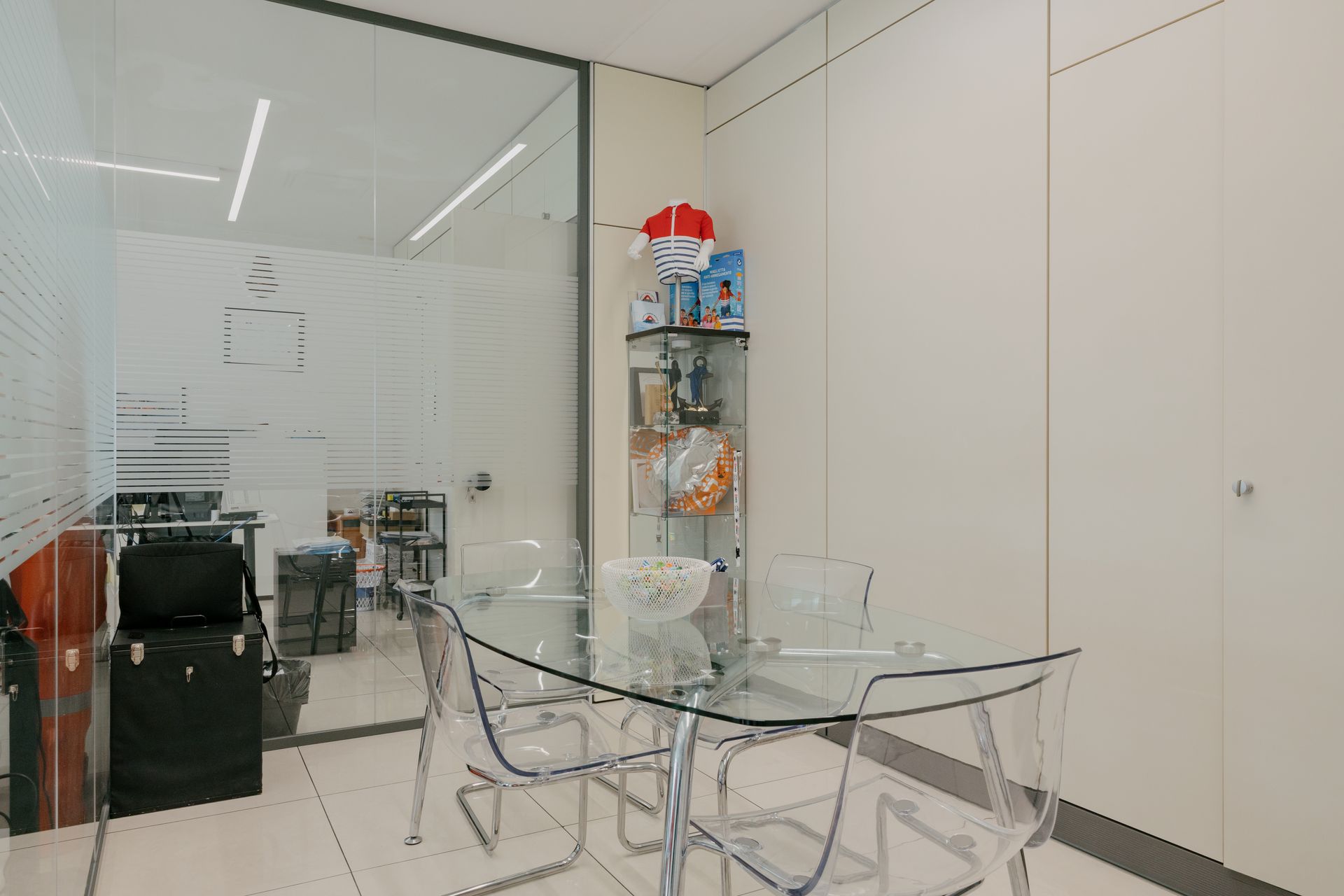 Small glass meeting room with a clear table, transparent chairs, and a shelf against white walls.