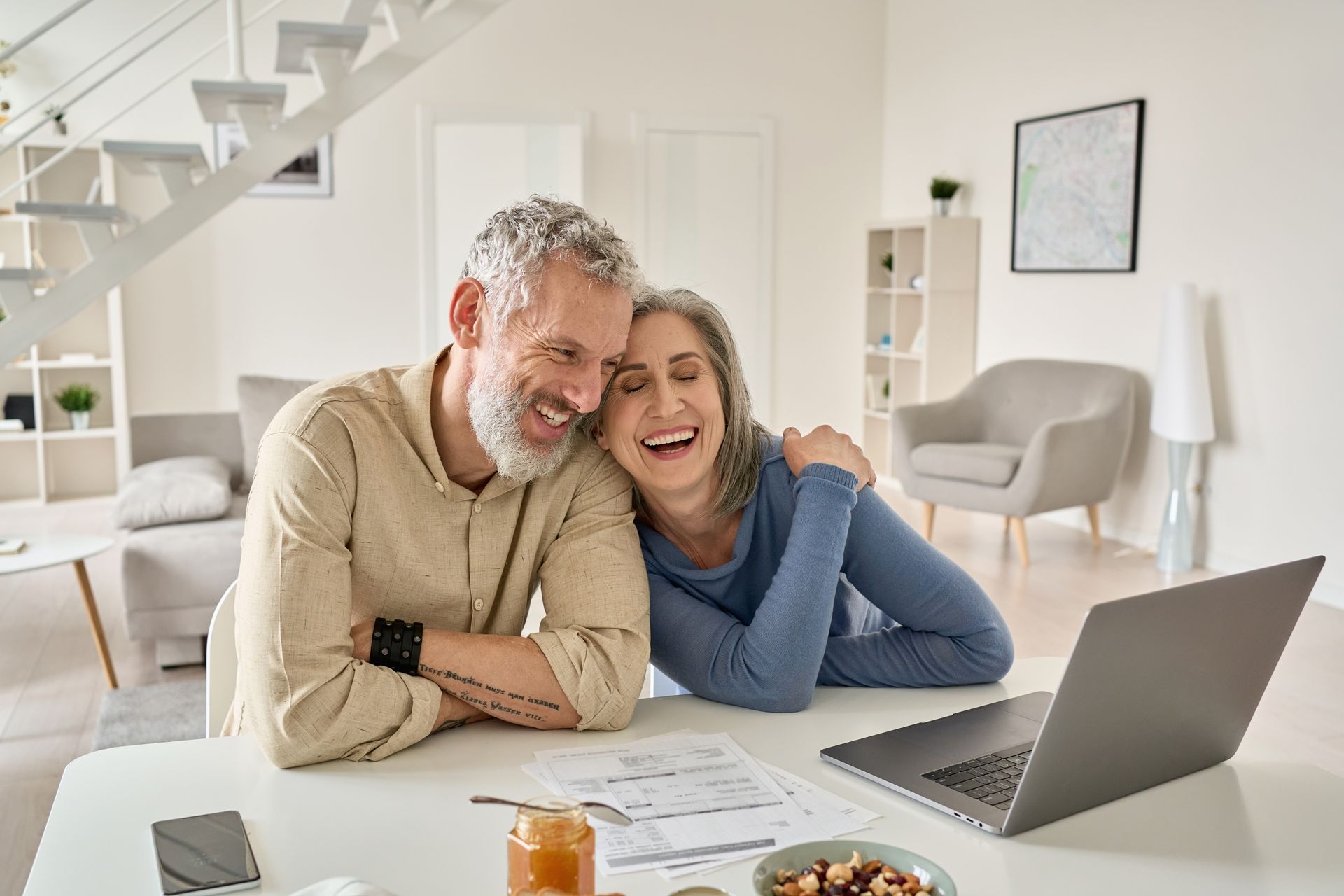 Couple laughing together at a table with a laptop, bills, and snacks in a bright living room.
