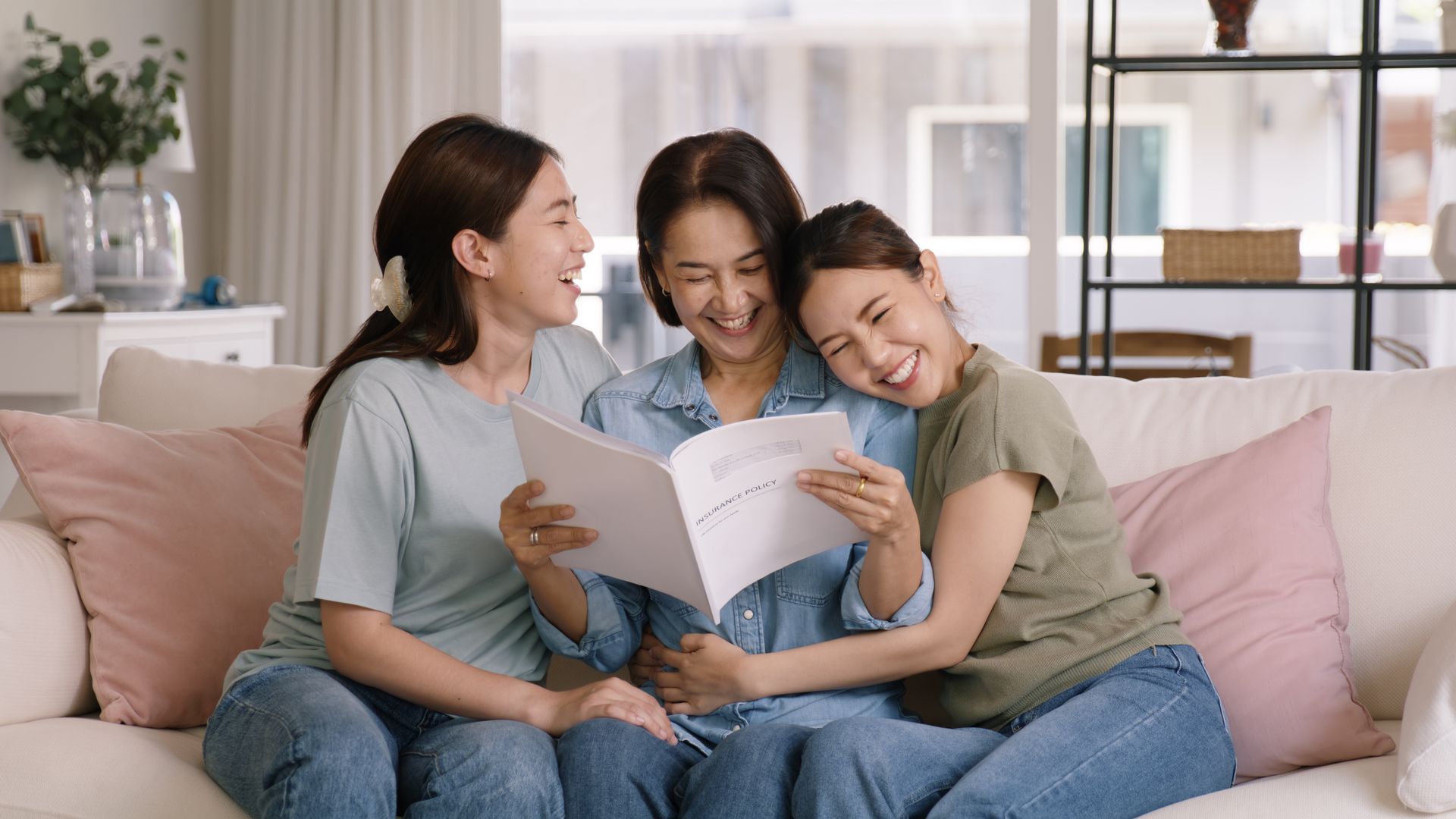 Three women laughing, looking at a photo album on a white couch in a living room.