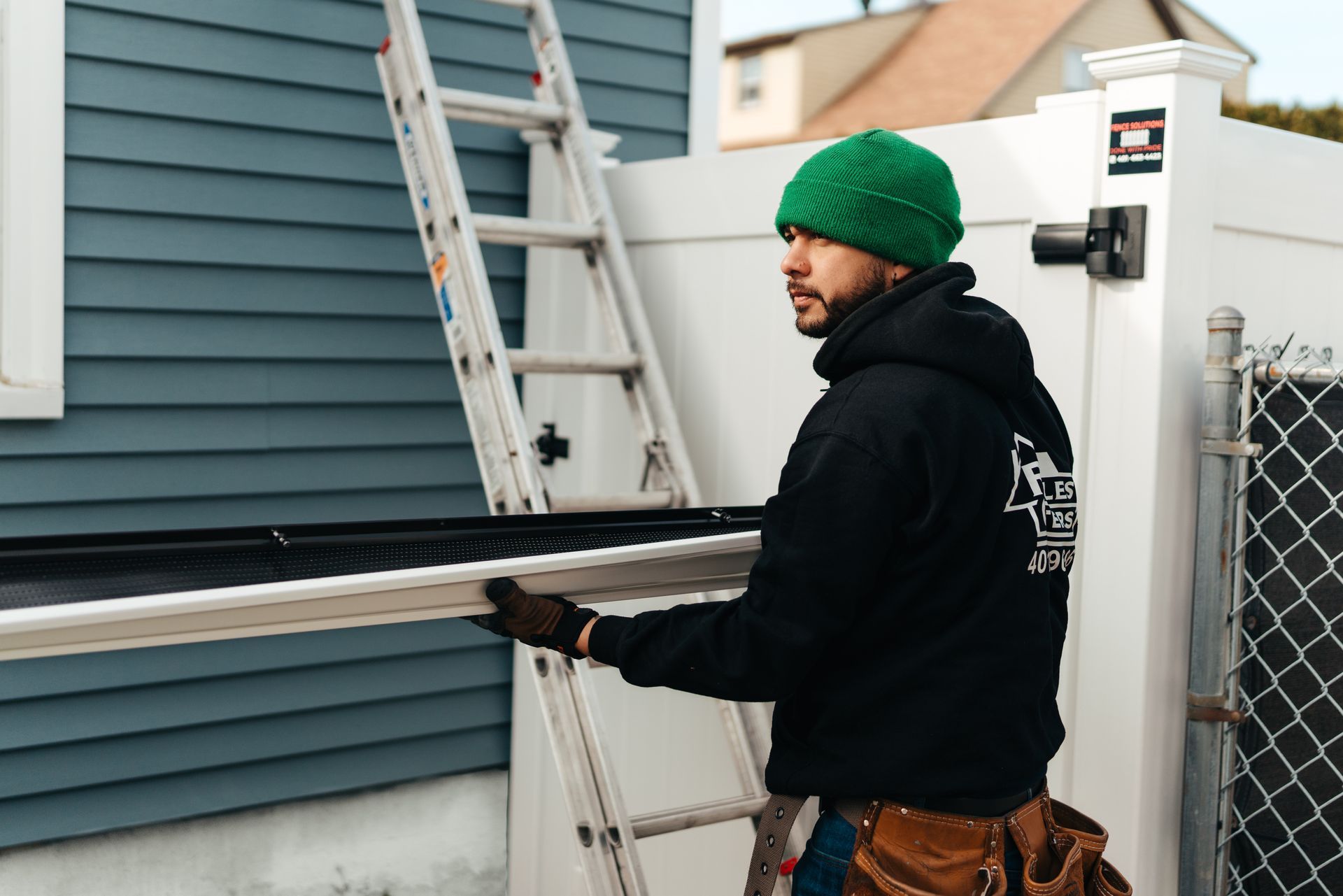 A man in a green hat is standing on a ladder next to a house.