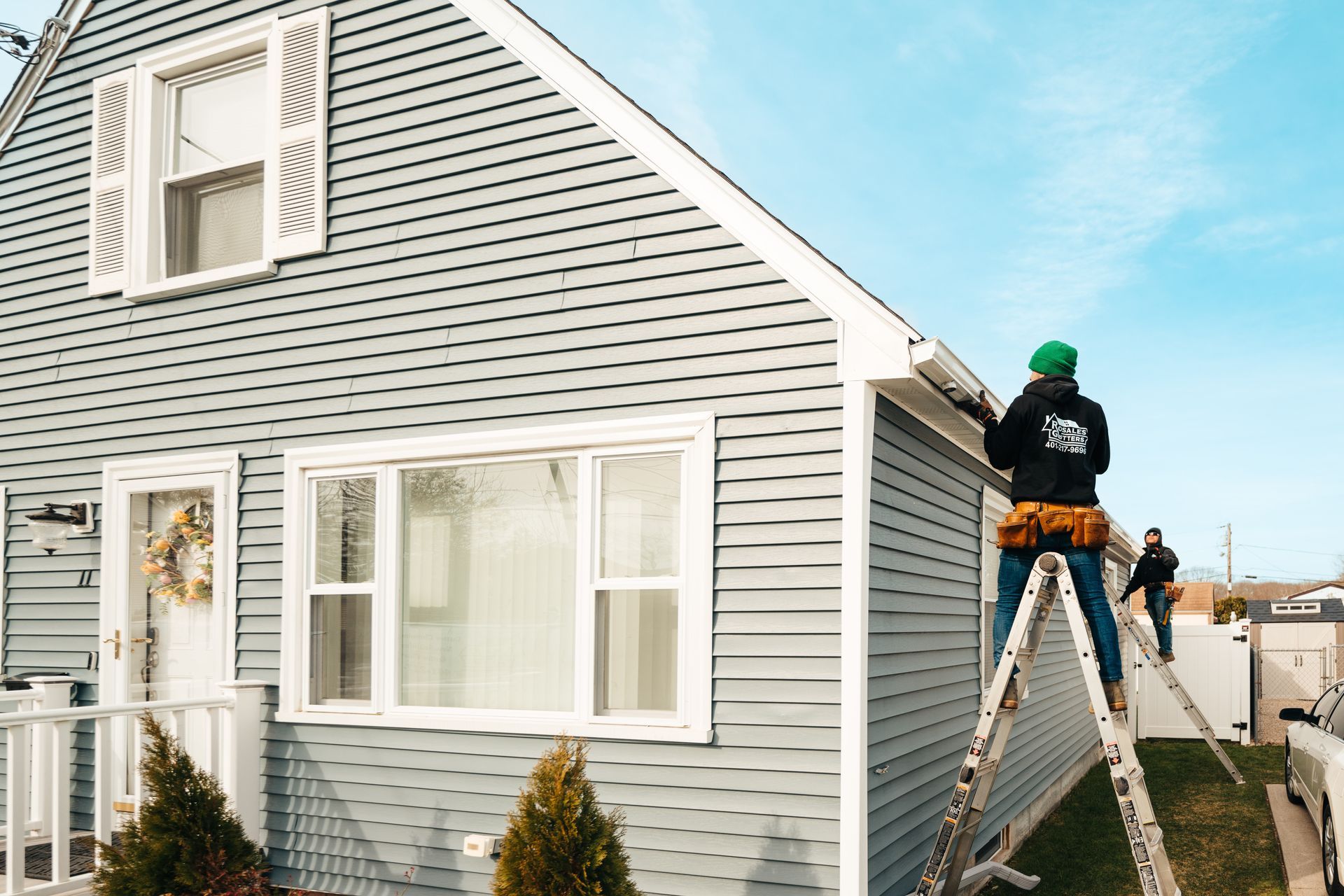 A man is standing on a ladder fixing a gutter on the side of a house.