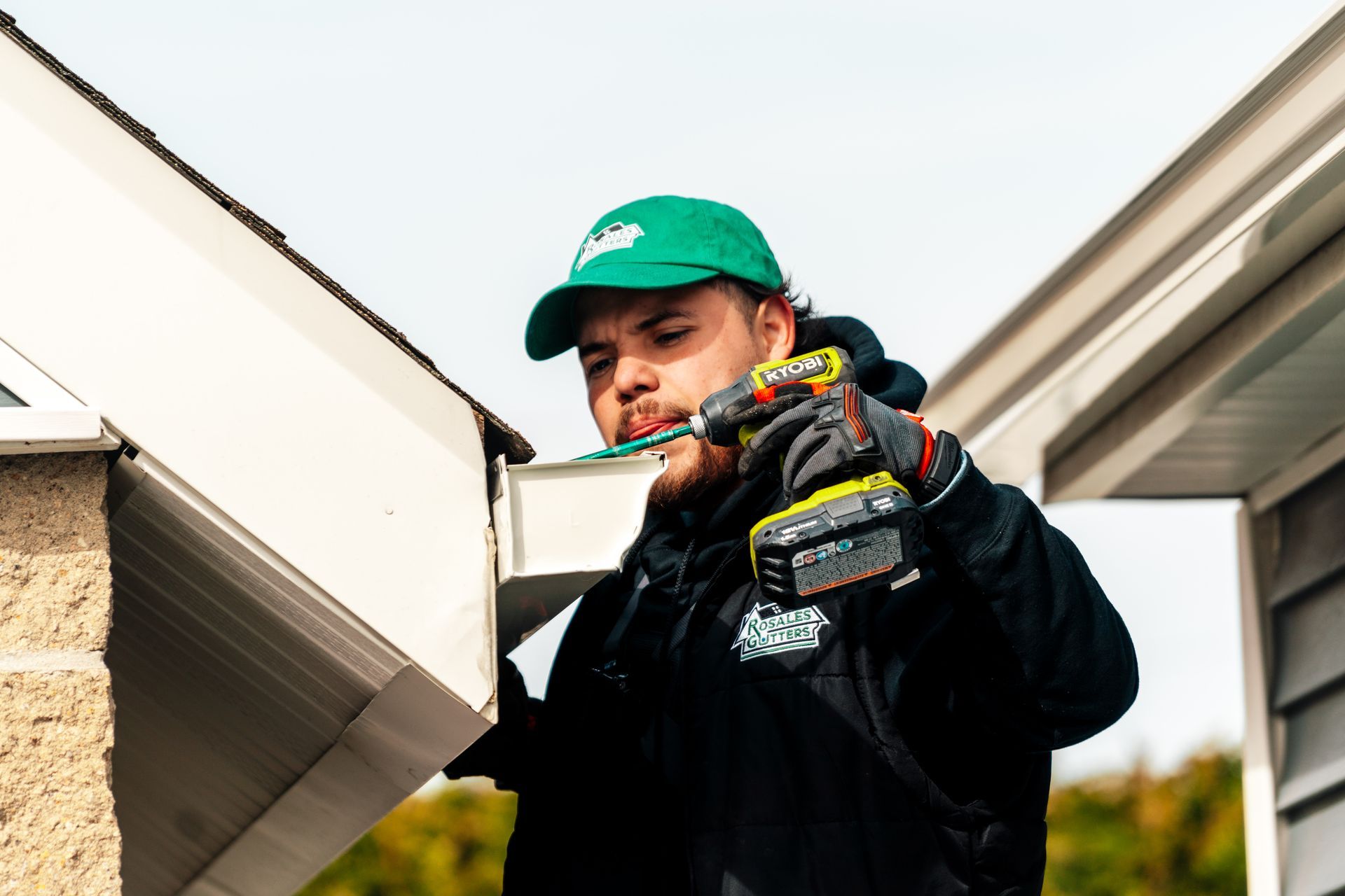 A man is using a drill to fix a gutter on the side of a house.