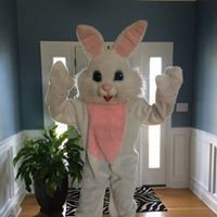 Person in a white bunny costume with pink ears and belly, waving in a home entryway.