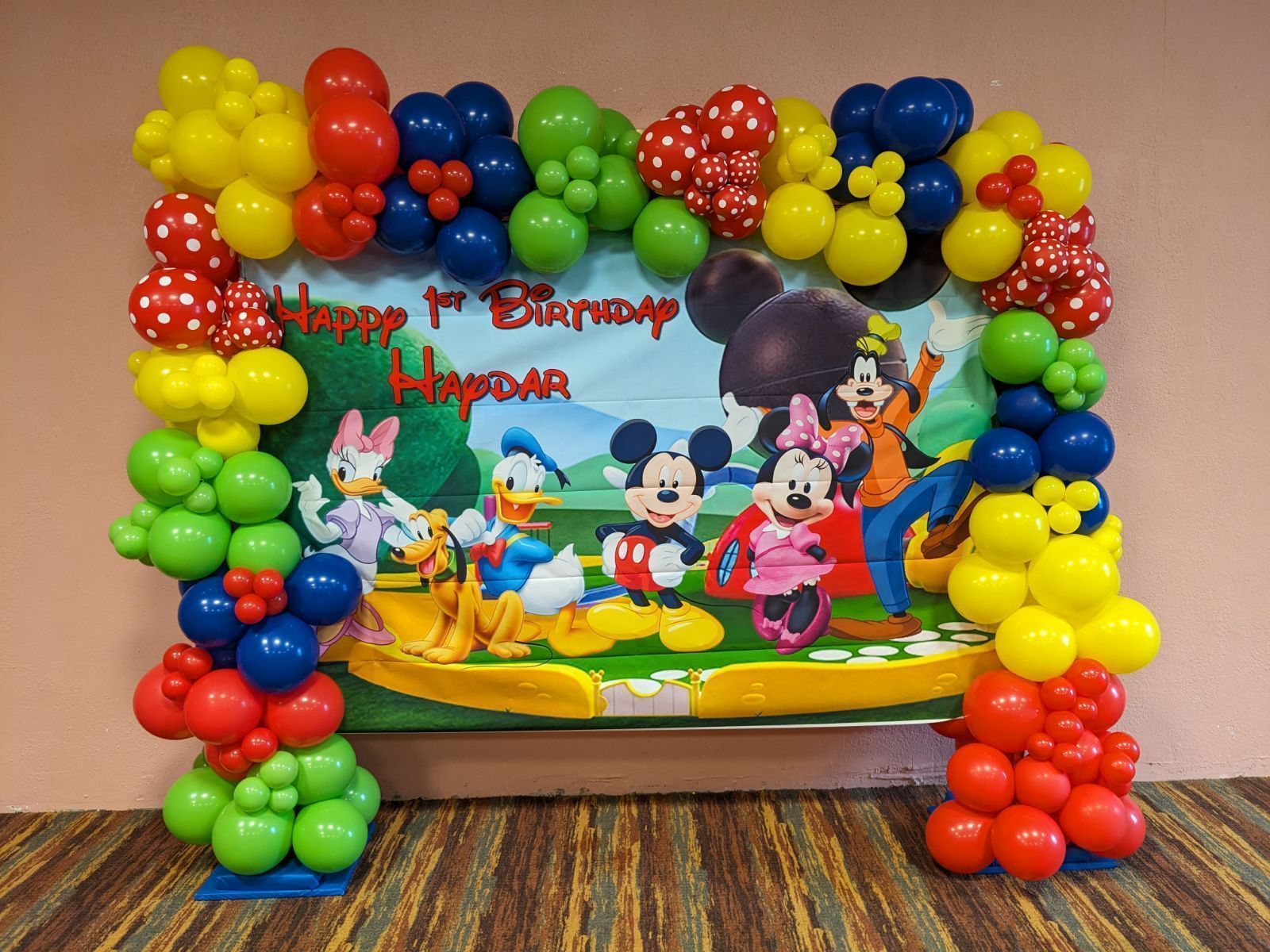 Mickey Mouse party backdrop framed by colorful balloon clusters on a wooden table, with cartoon characters in center