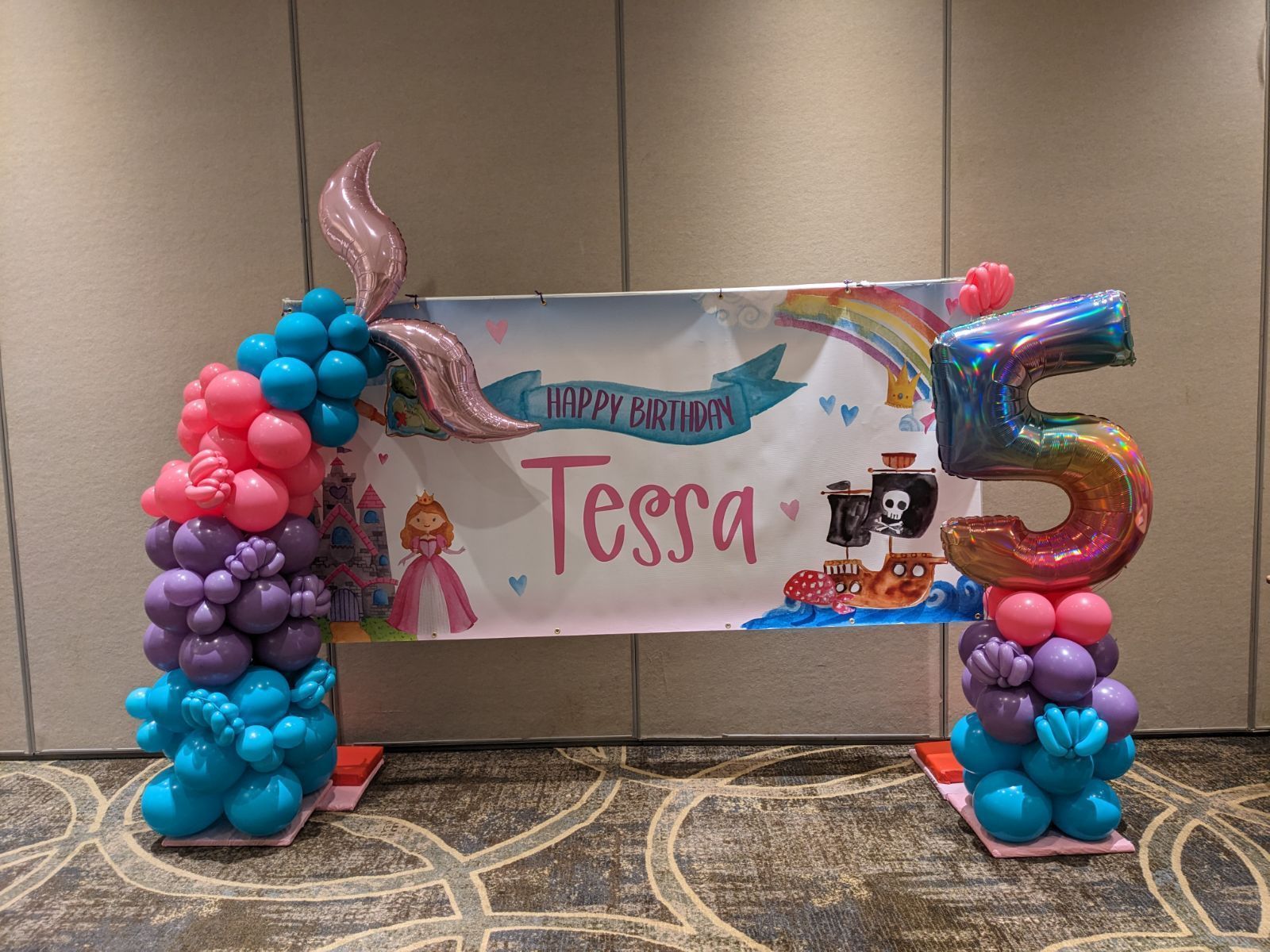 Birthday balloon arch with “Tessa” banner, pink and blue balloons, and a large number 5 in a hotel lobby