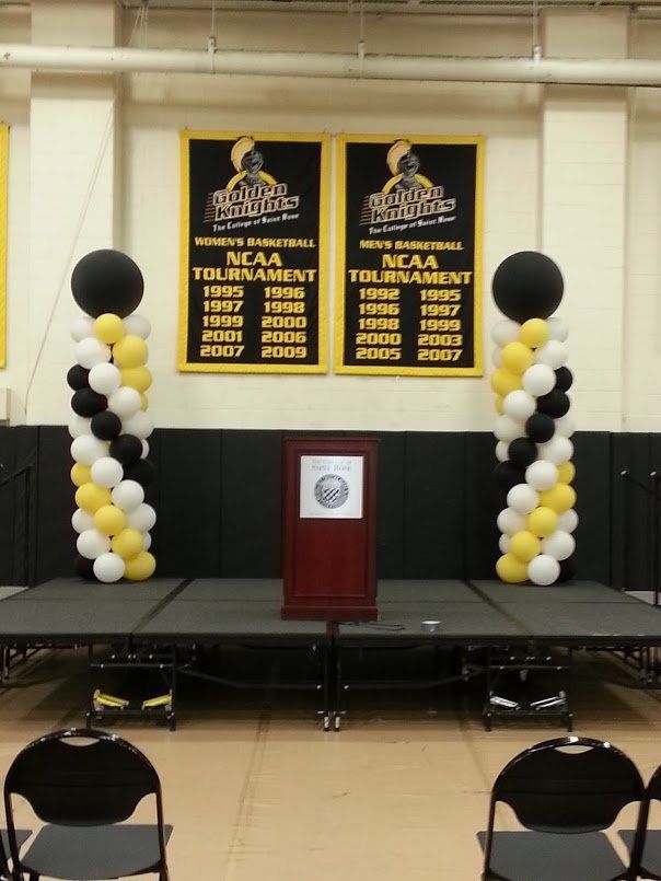 Stage with podium and black, gold, and white balloon columns beneath two NCAA banners
