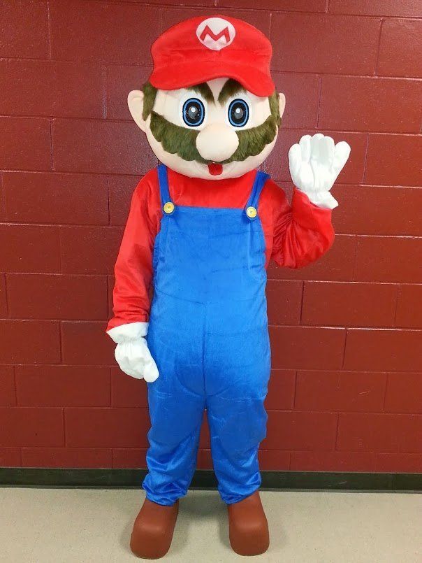 Mario mascot in red hat and blue overalls waving in front of a red brick wall