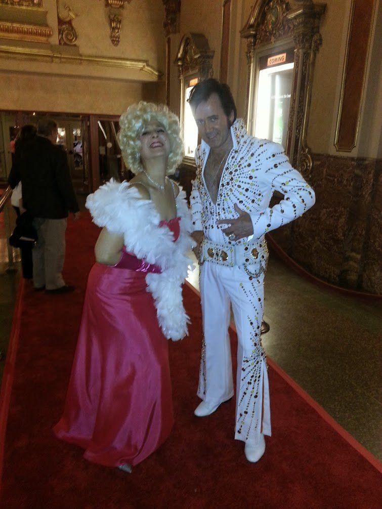Two costumed people posing in a hallway, one in a pink dress and boa, the other in a white rhinestone Elvis suit.