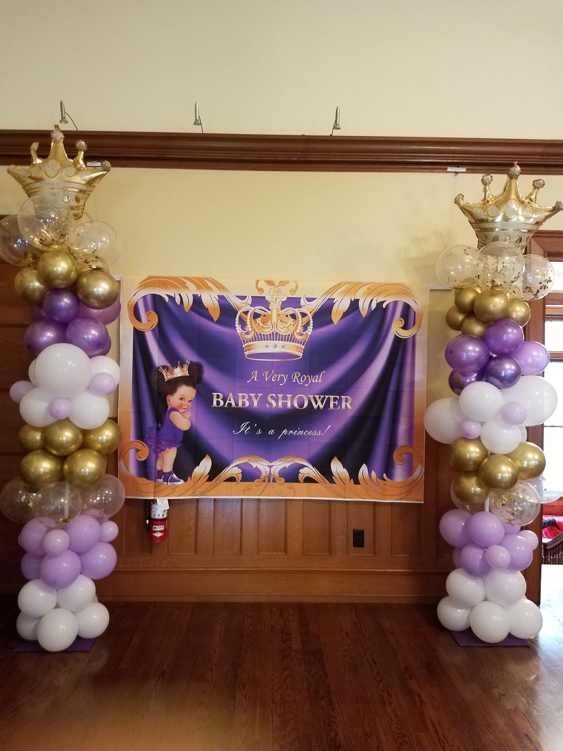 Purple-and-white baby shower backdrop with balloon columns and a central banner on a wood floor