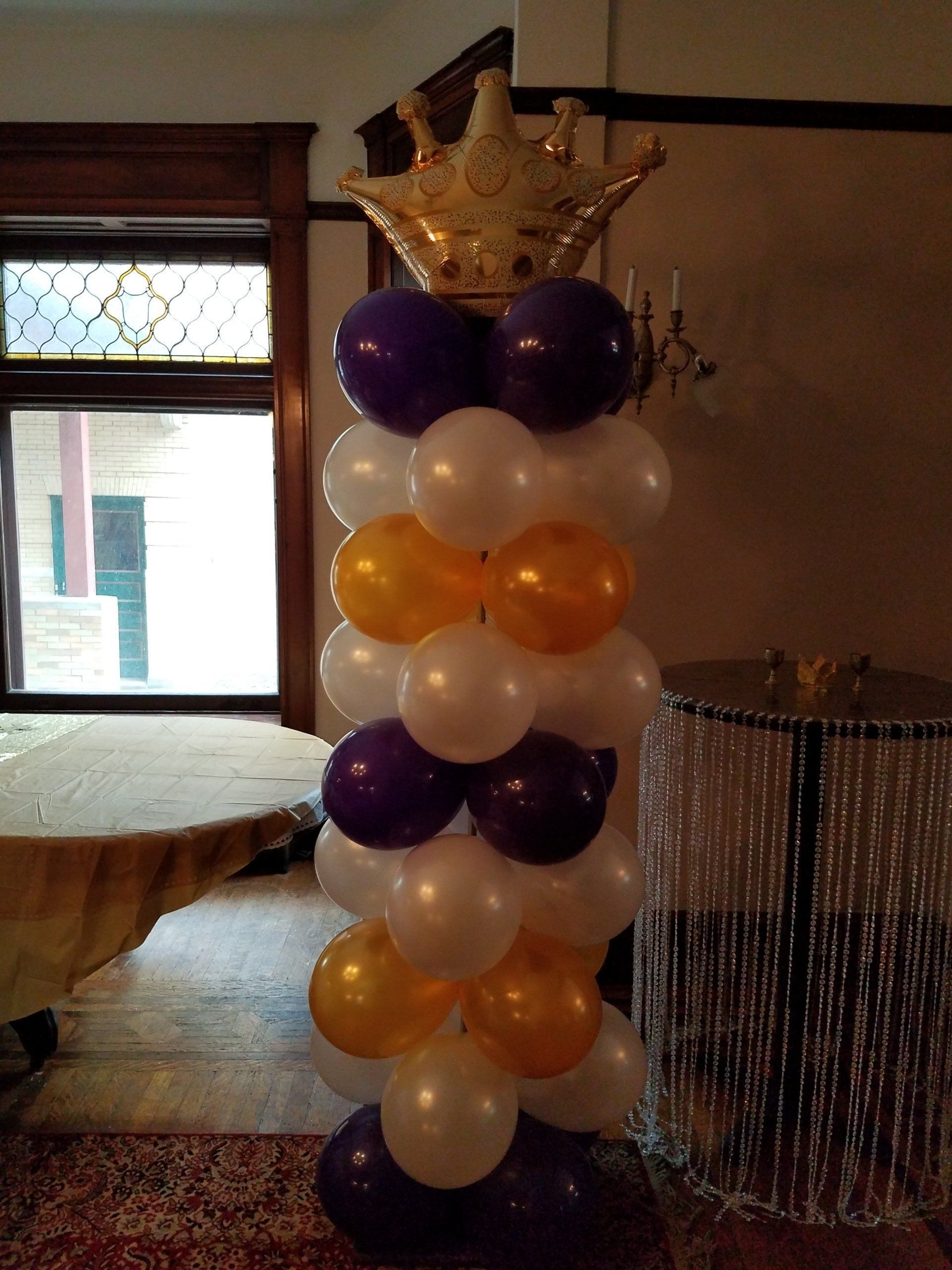 Balloon column with purple, white, and orange balloons topped by a gold crown in an indoor room