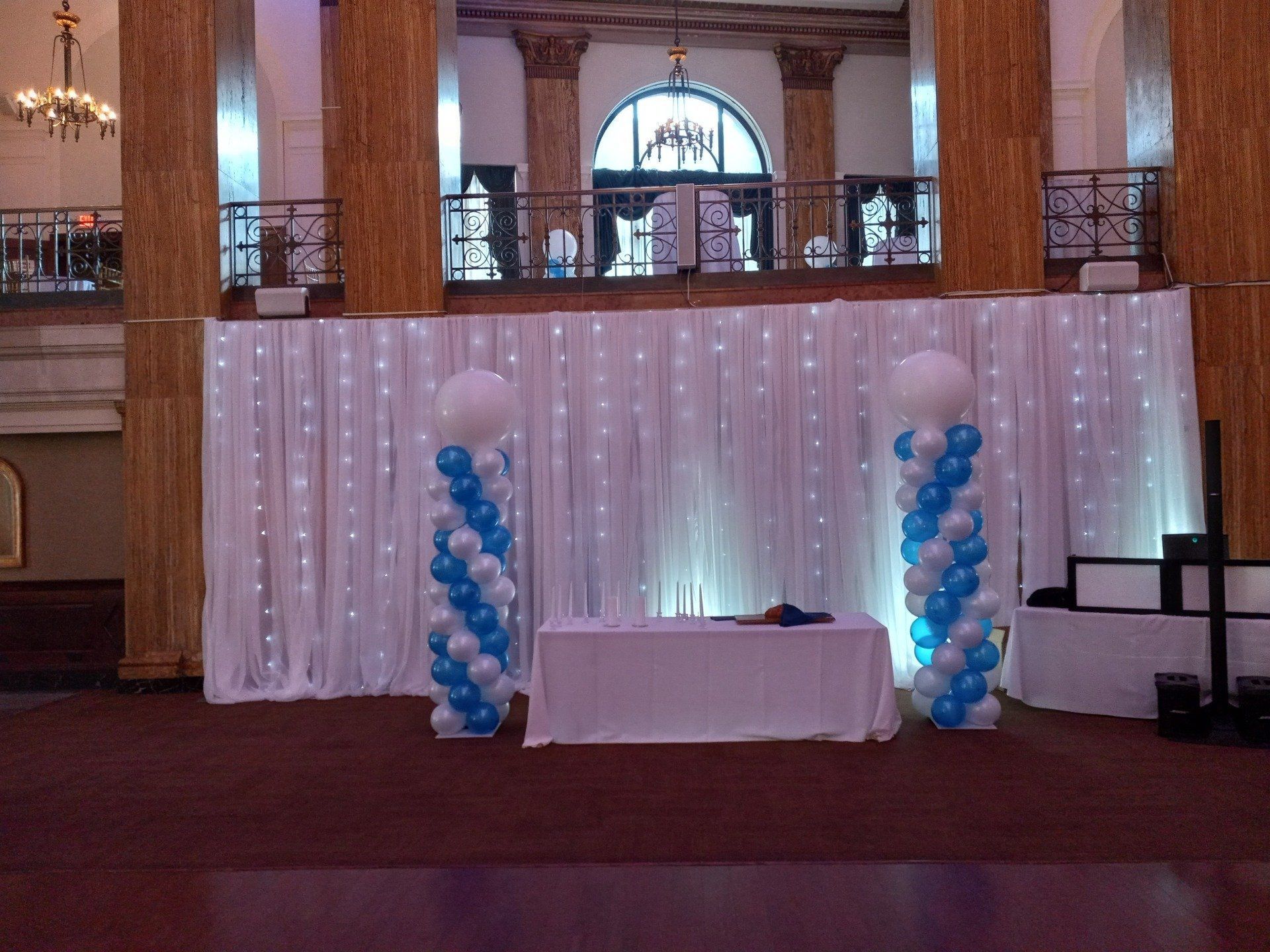 Decorated banquet hall stage with white drapery, blue balloon columns, and a small table centered in front