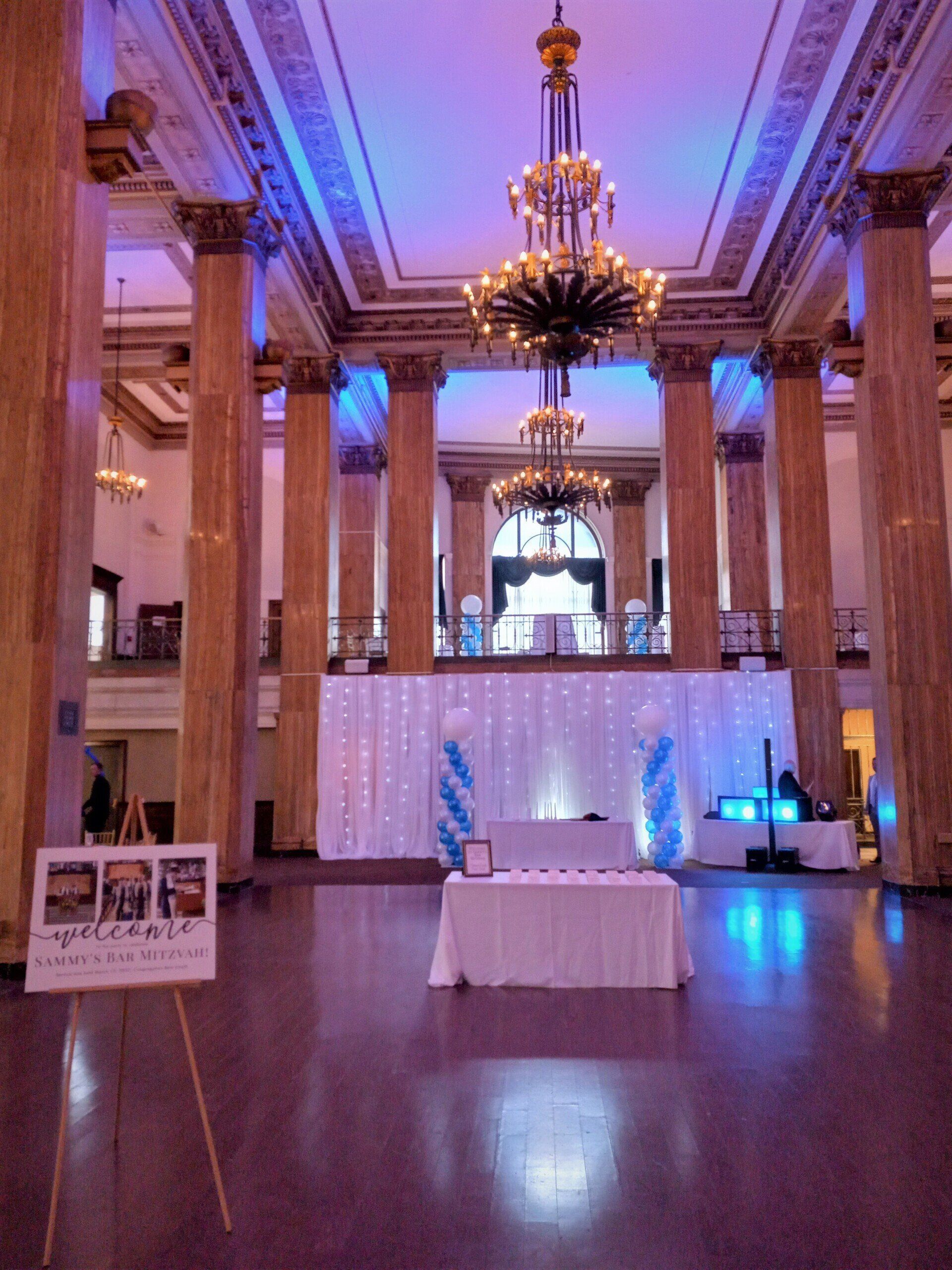 Grand hall with marble columns, chandeliers, and a small display table lit in blue and purple.