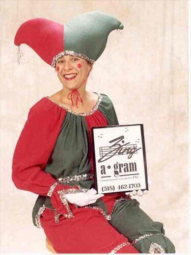 Person in red and green jester costume smiling and holding a sign reading “a gran”