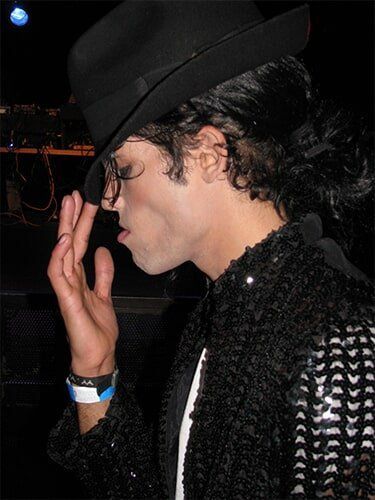 Person in a black hat and sequined jacket, profile view, touching their face in a dark setting.