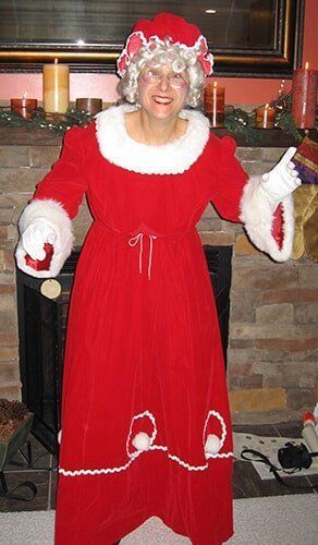 Person in a red Santa dress and white wig stands by a fireplace holding a candle.