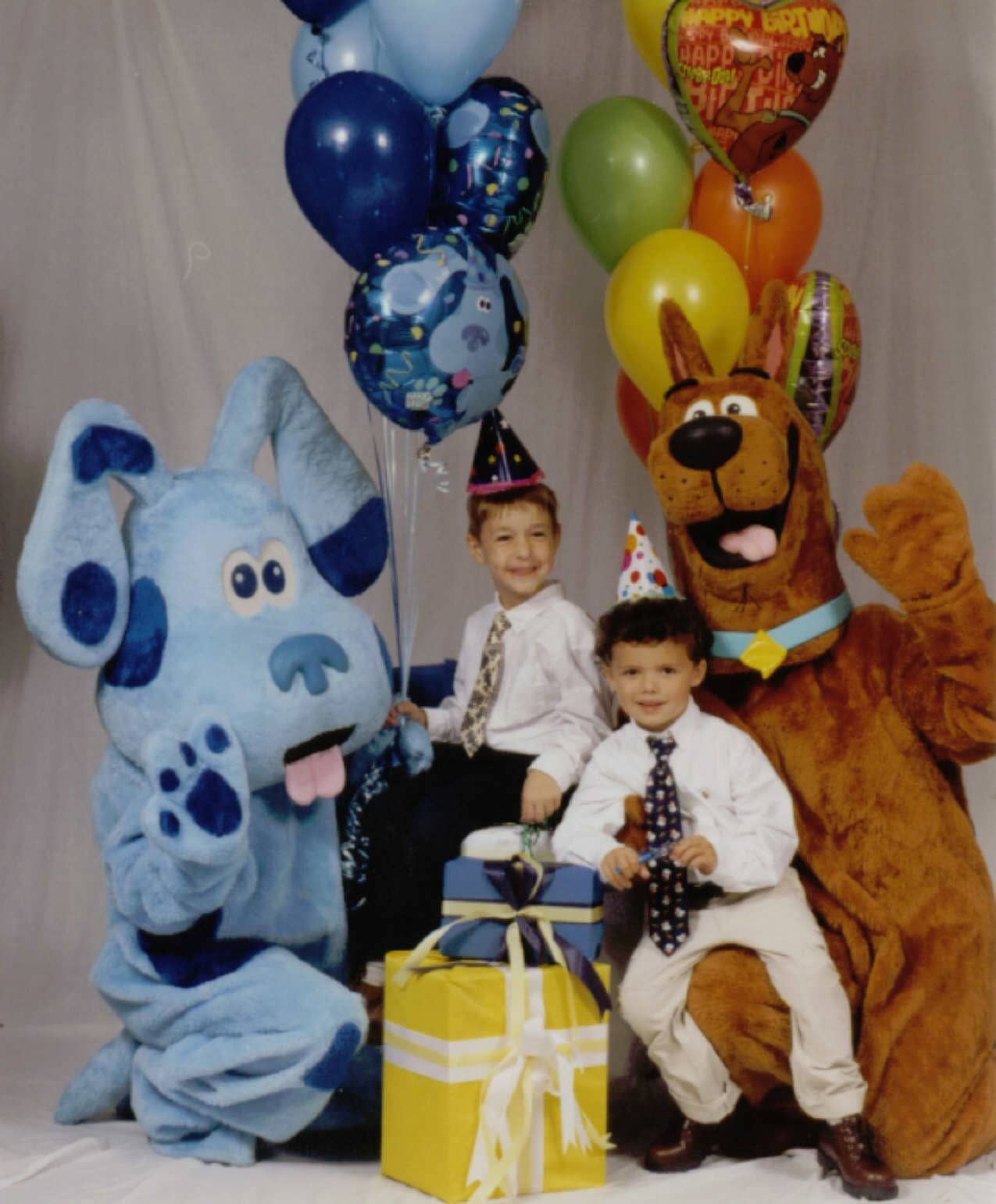 Two children with party hats seated by gift boxes, flanked by blue and brown cartoon mascots and colorful balloons.