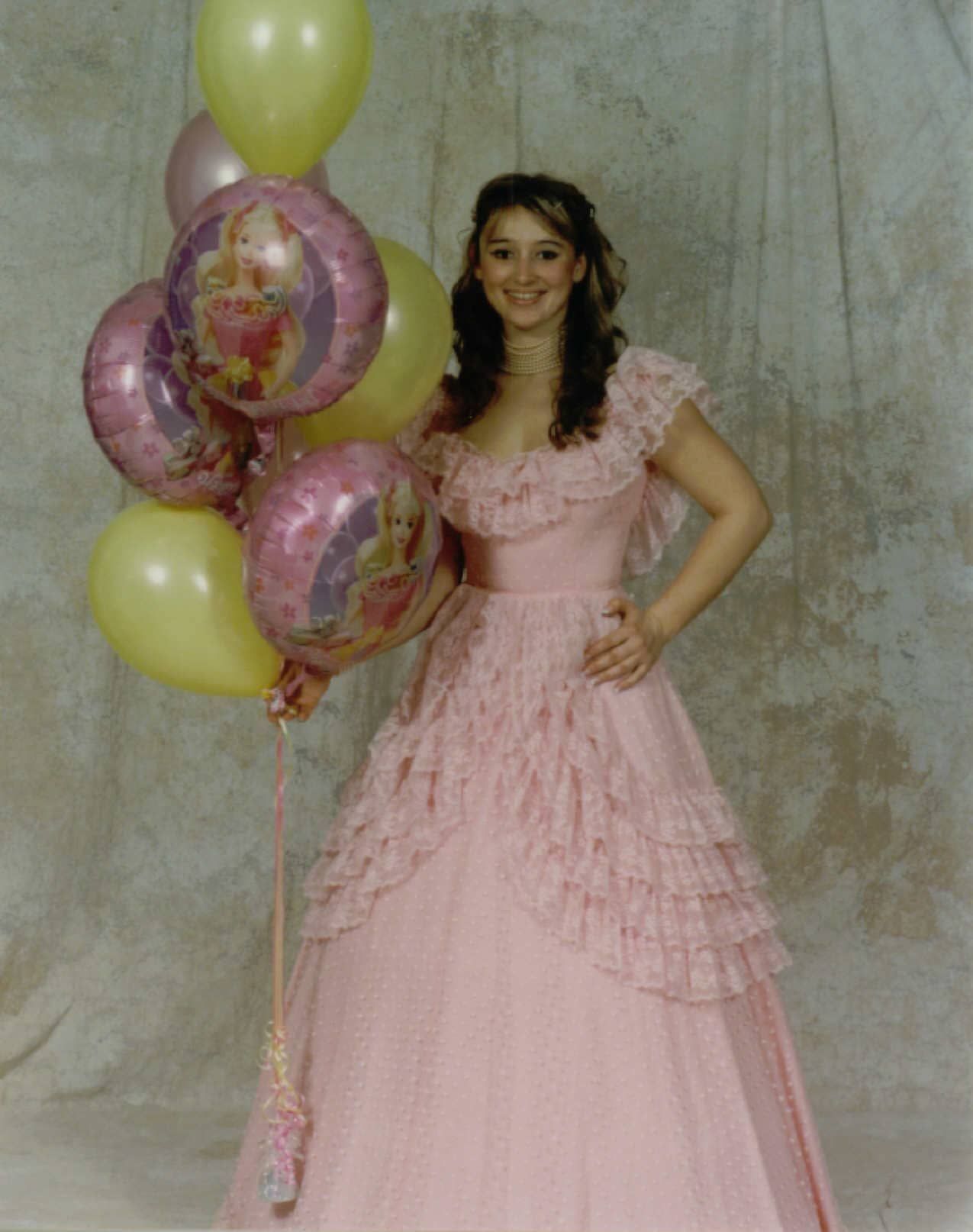 Person in a pink dress standing with balloons against a plain backdrop.