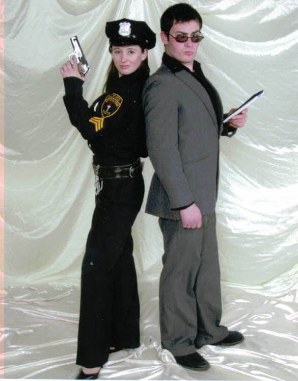 Two people in dark uniforms pose back-to-back, each holding a prop gun, against a white backdrop.