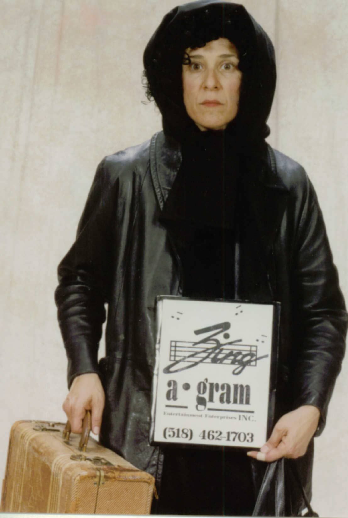 Person in black hooded coat holding a large sign and standing beside a suitcase.