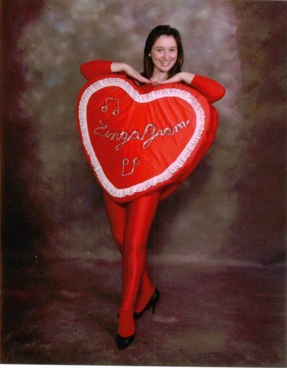 Person in a red heart costume posing against a studio backdrop, smiling with arms resting on the heart.