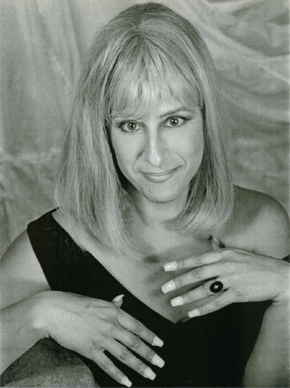 Blonde person in a black dress posing with hands near chest against a draped backdrop