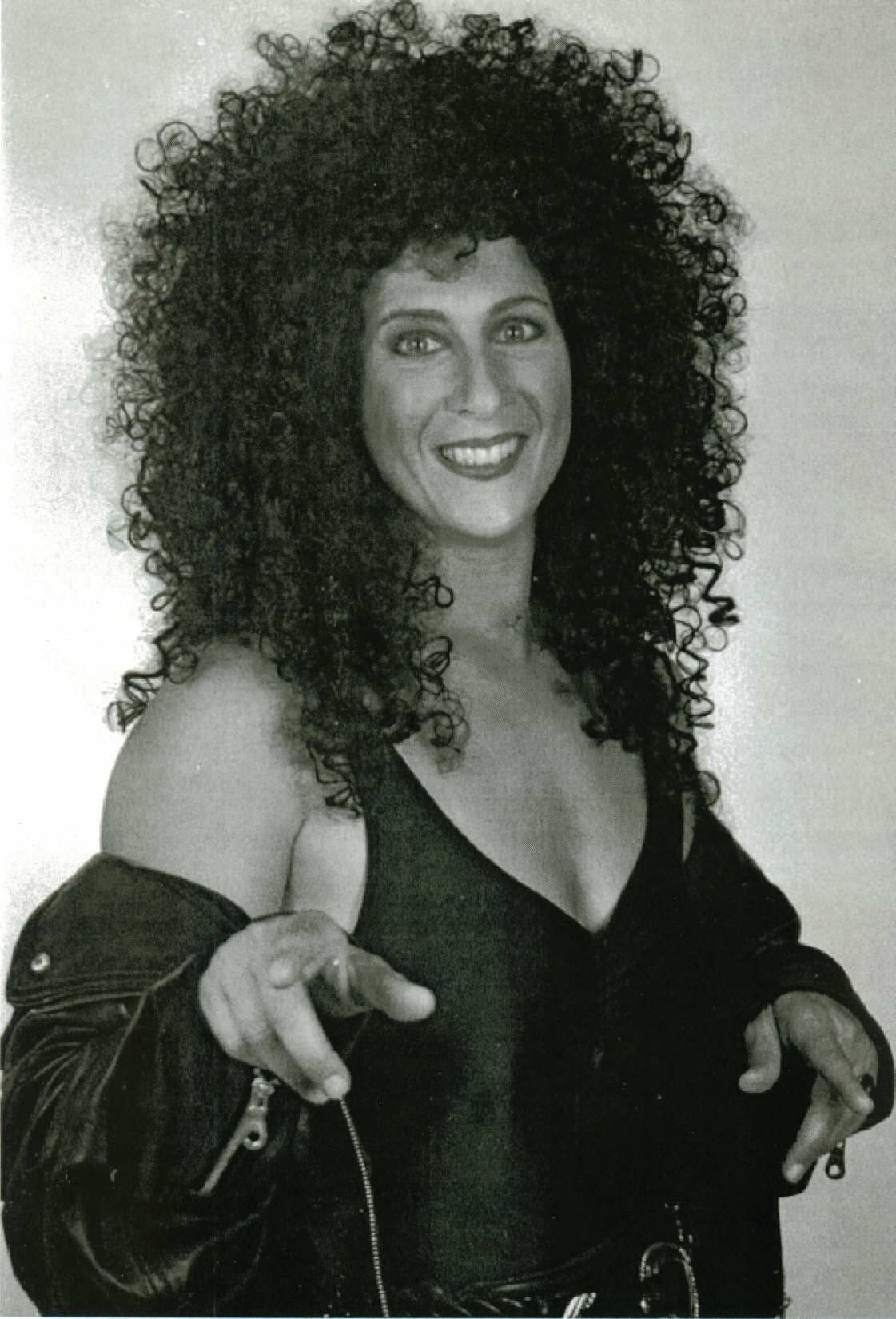 Smiling person with curly hair in black outfit, holding hands up against a plain background