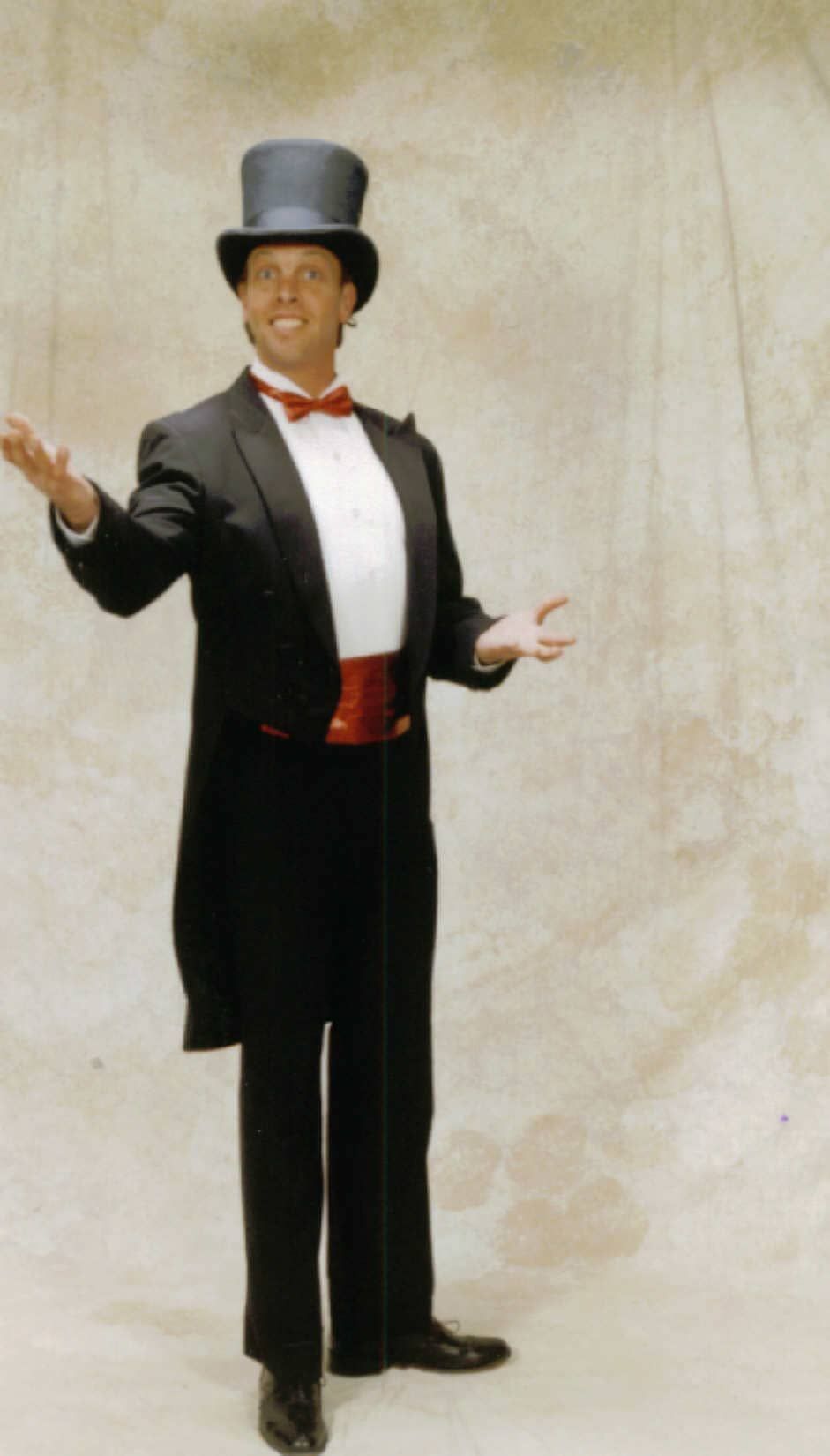 A man in a black tuxedo and top hat stands with arms outstretched against a plain backdrop.