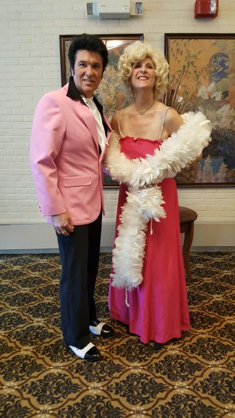 Two people in formal costumes pose indoors; one wears a pink suit, the other a pink dress with a white feather boa.