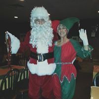 Santa and an elf posing together indoors, both waving at the camera.