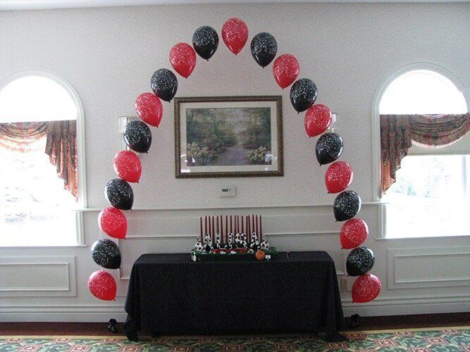 Black and red balloon arch framing a framed picture above a black-draped table in a bright room