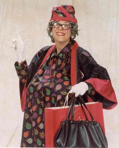 Woman in a colorful outfit and hat, smiling and holding shopping bags against a plain background