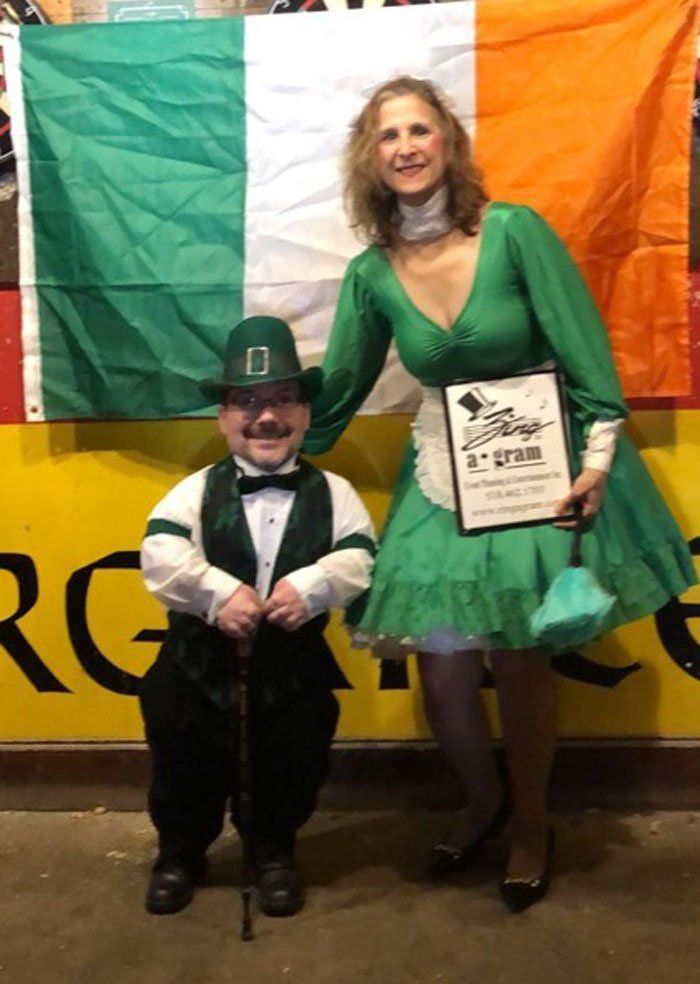 Two people in green outfits pose in front of an Irish flag, one holding a plaque.