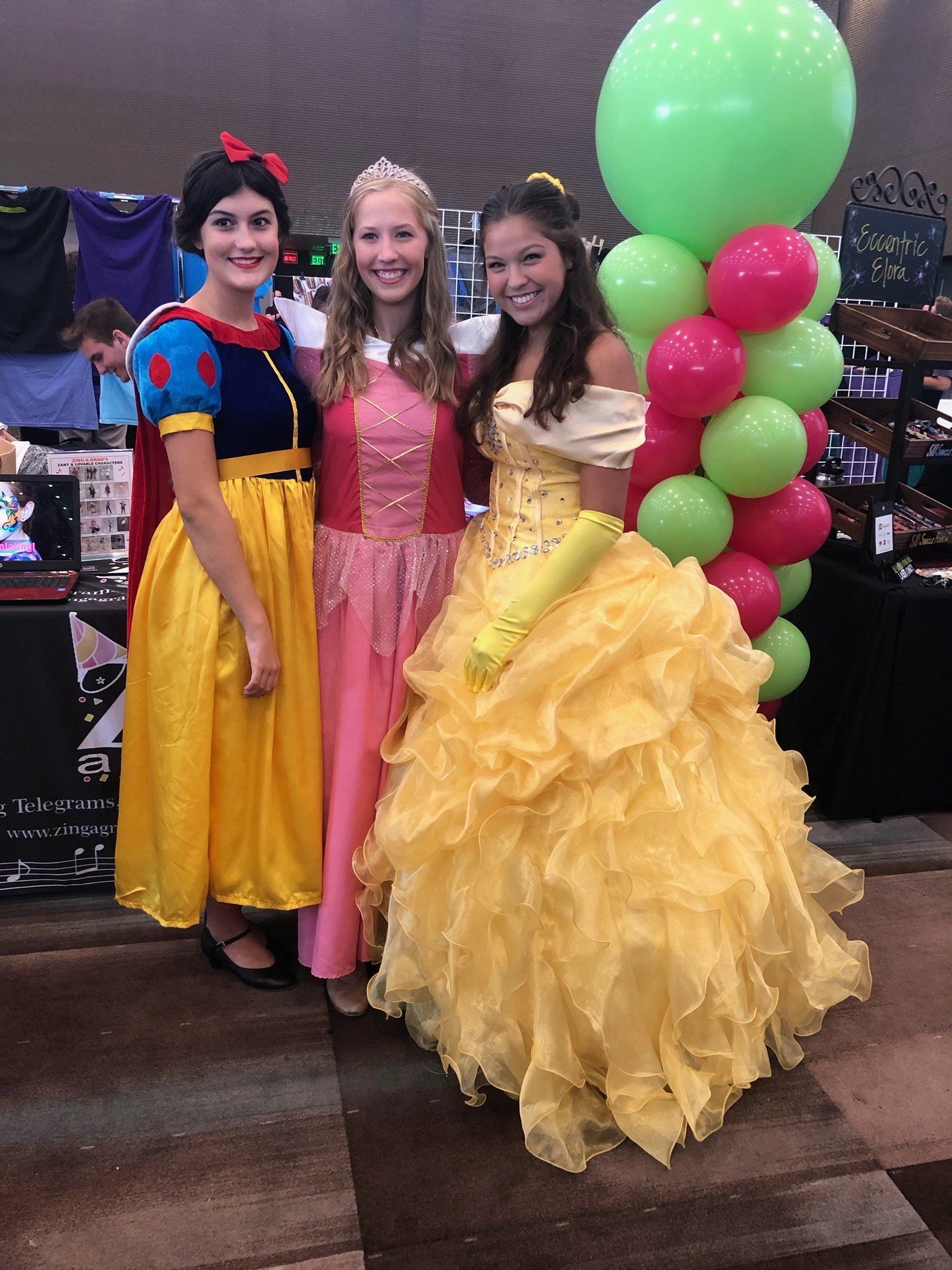 Three children in colorful princess dresses posing indoors beside a tall green and pink balloon arrangement.