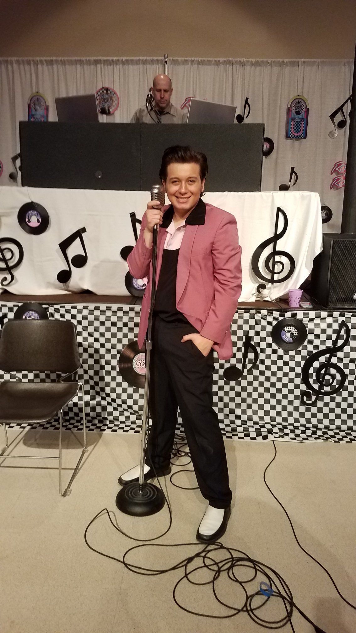 Child singing into a microphone on a small stage with music-note decorations and a DJ booth behind.