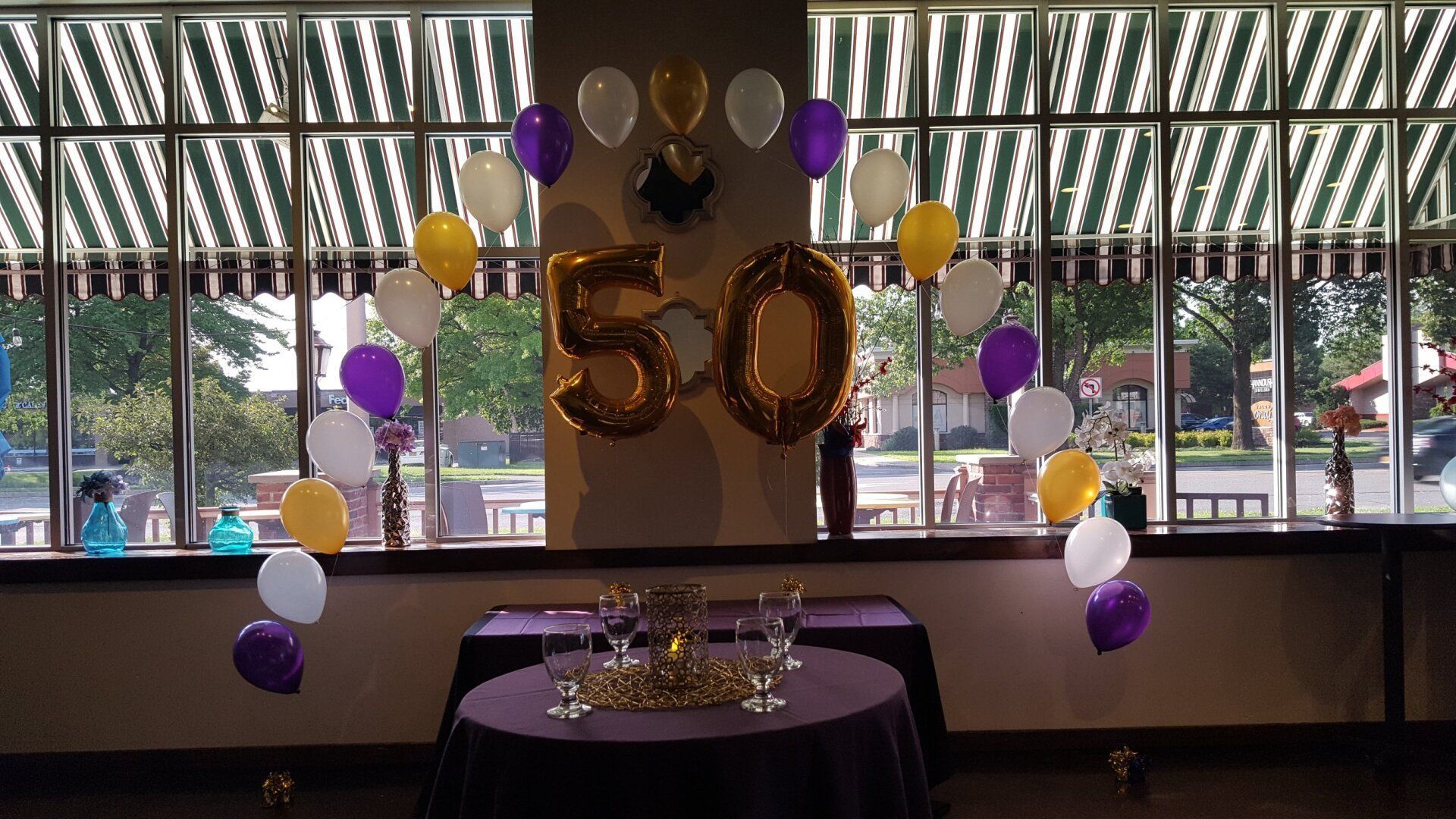 Gold “50” balloon display with purple, gold, and white balloons at a decorated party table by windows