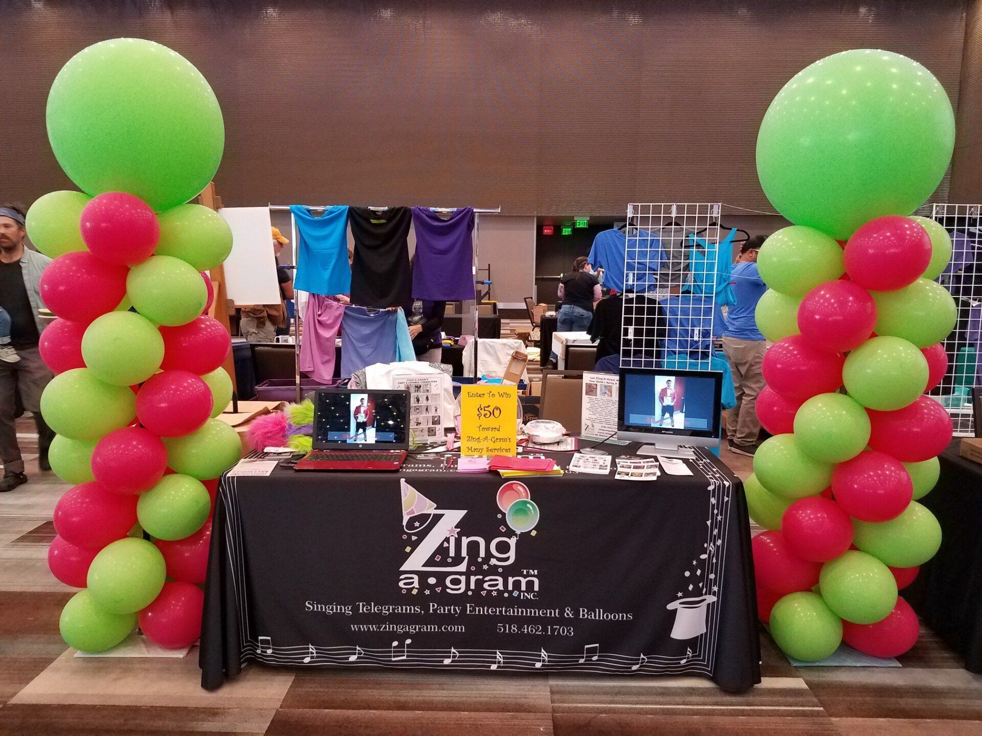 Vendor booth with black tablecloth, balloon columns, hanging shirts, and electronics on display.